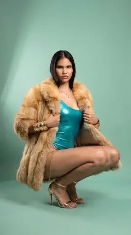 Glamorous woman in a teal bodysuit and faux fur coat posing in a studio setting.