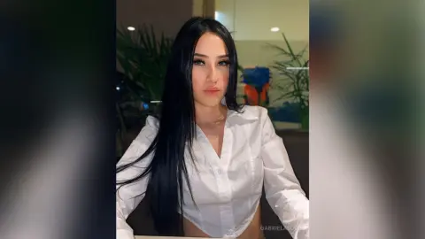Young woman with long black hair wearing a white button-down shirt in a modern indoor setting.