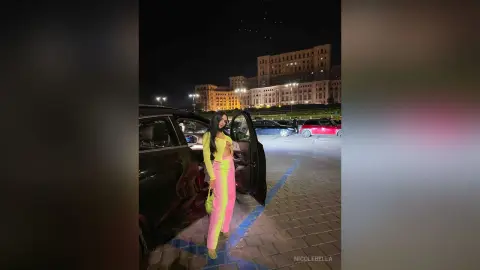 NicoleBella stands next to an open car door wearing a bright yellow crop top and pink leggings or trousers at night.