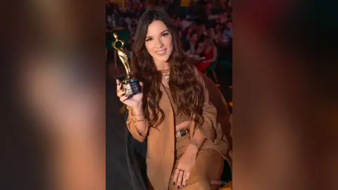Rebecca smiles while holding a gold award trophy, wearing a brown crop top and matching trousers.