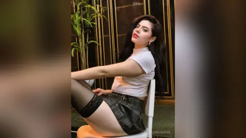 Young woman in white crop top and plaid skirt sitting in a modern indoor setting with a moody aesthetic.