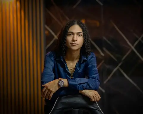 KylePresly wears a dark blue button-down shirt and a gold necklace while leaning forward against a dark chair.