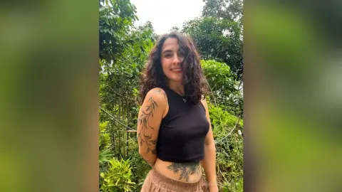 RossiNoleen smiles while wearing a black tank top that shows her floral arm and stomach tattoos against a lush green forest background.