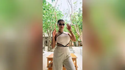 Stylish woman in a white halter jumpsuit and sunglasses standing in a lush green outdoor garden setting.