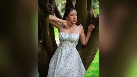 IsabellaYork — Young woman in a white floral sundress leaning against a tree in a lush green park with a dreamy mood.