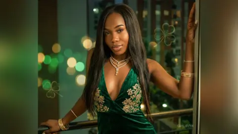 Elegant woman in a green velvet evening gown posing on a balcony with glowing city lights at night.