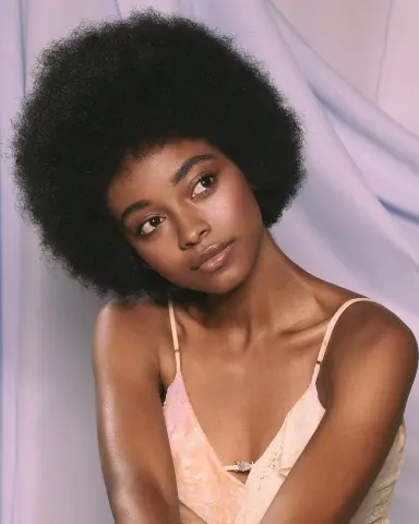 Young Black woman with afro hairstyle wearing a peach camisole against a soft lavender fabric background.