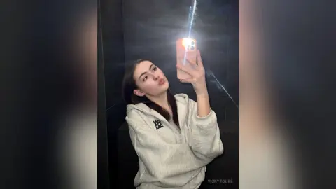 VickyToure takes a mirror selfie wearing a light grey hooded sweatshirt, showing her face and long dark hair against a dark background.