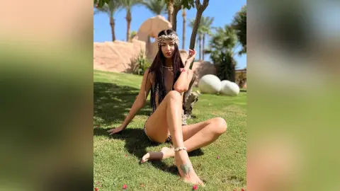 Woman in bohemian crochet outfit sitting on green grass in a sunny tropical garden setting.