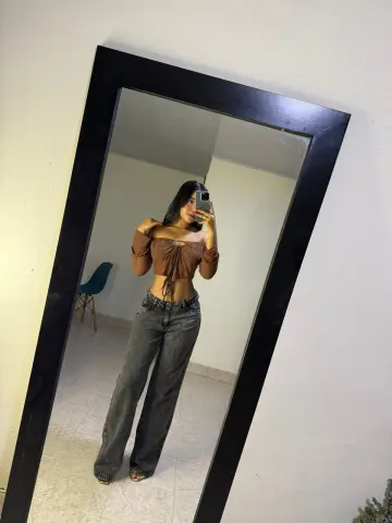Young woman in a brown off-the-shoulder crop top and grey wide-leg jeans taking a mirror selfie in a minimalist room.