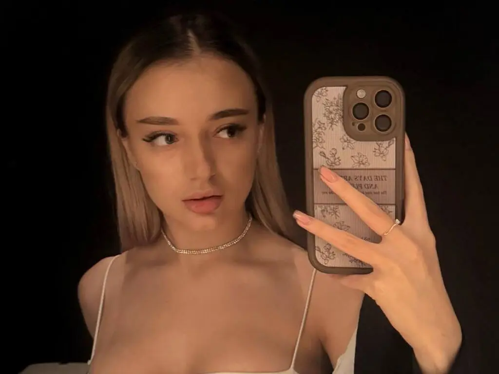 Young woman in a white camisole taking a moody selfie in a dark room with a patterned phone case.