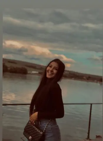 Smiling young woman in a black long-sleeve top and jeans standing by a lake under a cloudy sunset sky.