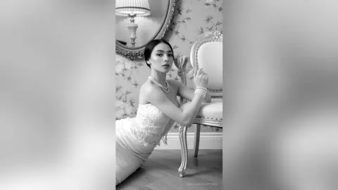 Elegant woman in a white lace gown and pearl necklace sitting in a classic vintage room, black and white portrait.