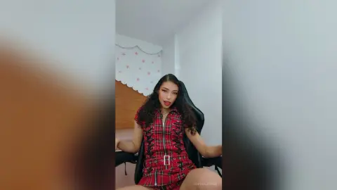 Young woman with curly hair wearing a red plaid romper sitting in a gaming chair in a bright bedroom.