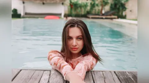 StacyHarland leans over a wooden deck edge with her long brown hair falling forward against a bright blue swimming pool.
