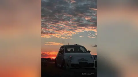 White Fiat 500 parked under a dramatic orange and pink sunset sky during golden hour.