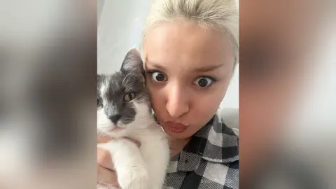 BarbaraGreen pouts her lips while holding a grey and white cat close to her face in a close-up selfie.