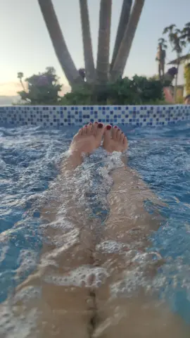 Relaxing feet dipping into a sparkling blue swimming pool at a tropical resort during sunset.