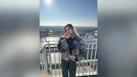 ValerieGracy stands on a balcony wearing a grey fur coat and dark trousers against a bright sky and city river view.