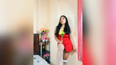 Young woman in a red mini dress holding a yellow flower in a bright, cozy bedroom setting.