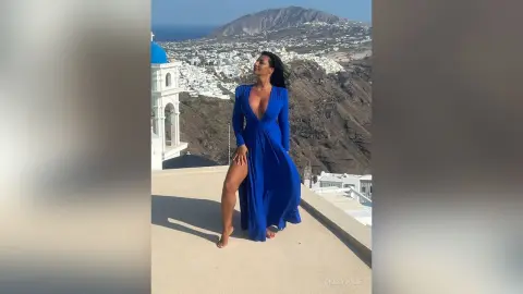 MissyJolie stands on a white terrace overlooking a coastal town, wearing a long, deep-V blue dress that shows her bare legs.