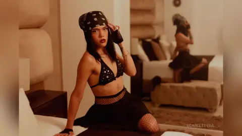 Confident woman in a black bikini top, bandana, and mesh skirt posing in a warm, moody bedroom setting.