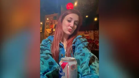 Woman in a blue patterned denim jacket holding a beverage in a dimly lit, cozy indoor setting.