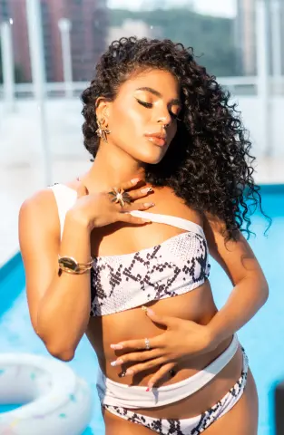 Smiling woman with curly hair in a snakeskin print bikini relaxing by a sunny blue swimming pool.