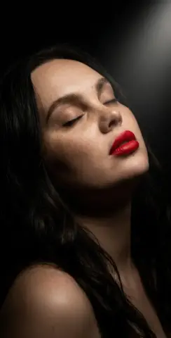 Close-up portrait of a woman with red lipstick and closed eyes in a moody, dark studio setting.