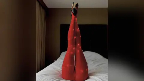 Woman in red bodysuit and high heels posing upside down on a white bed in a dimly lit bedroom.