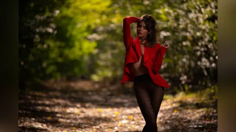 AnaMathers poses in a red open jacket and black tights against a sunlit, leafy forest background.