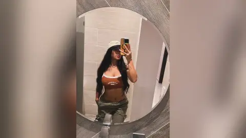 Young woman in a brown crop top and baggy trousers taking a casual mirror selfie in a modern bathroom.