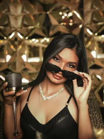 Glamorous woman in a black camisole and sunglasses holding a cocktail in a luxury gold setting.