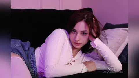 Young woman in a white long-sleeve top relaxing on a dark sofa with moody purple lighting.