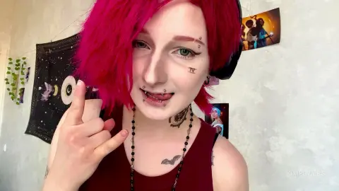 Woman with pink hair and facial piercings wearing a maroon tank top in a cozy bedroom setting.