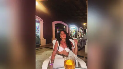 RossiNoleen smiles while sitting in a white chair wearing a white lace bra and white skirt, holding a glass of beer.