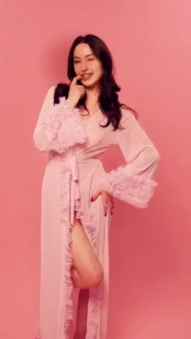 Smiling woman in a light pink ruffled silk robe posing against a soft pink background.