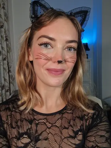 DaisyWrenUK smiles at the camera wearing black lace and cat ears with whiskers painted on her face.