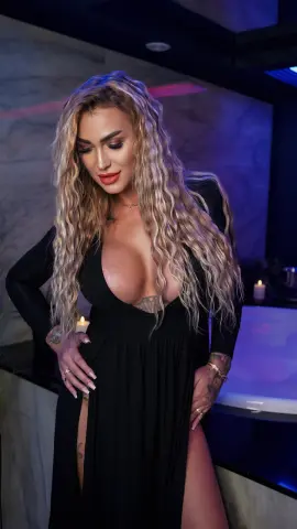 Blonde woman in a low-cut black dress posing in moody, blue-lit indoor setting.