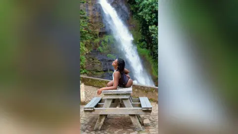 Curvy woman in a black swimsuit sitting on a wooden picnic table near a lush tropical waterfall.