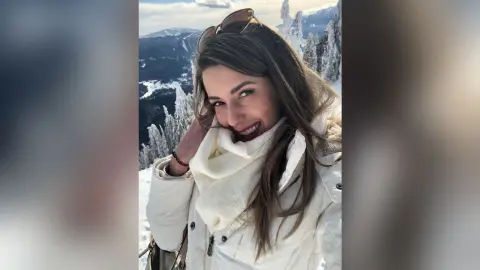 MontseFerrera smiles while wearing a white jacket and a thick white scarf in a snowy mountain setting.