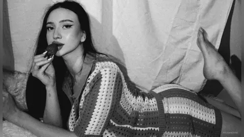 Moody black and white portrait of a woman in a patterned knit dress eating a strawberry on a white bed.