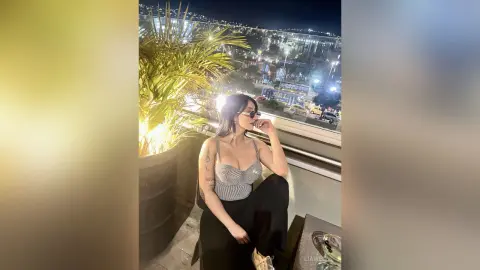 Young woman in a striped bodysuit relaxing on a rooftop balcony overlooking a glowing city skyline at night.