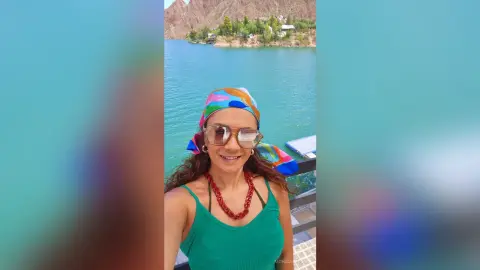 Smiling woman in a green tank top and colorful headscarf enjoying a sunny day by a turquoise mountain lake.