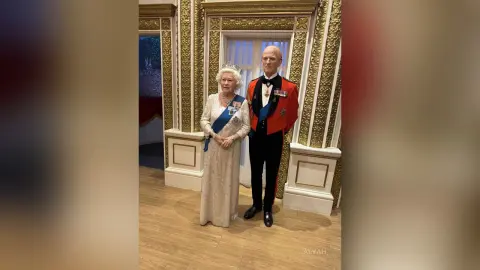 Queen Elizabeth II in a white gown and King Charles III in formal red military regalia standing in a grand palace.