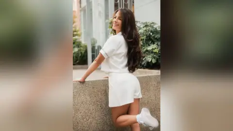 TefyRusso smiles while leaning against a stone wall, wearing a white short-sleeved top and matching white skirt with her long dark hair flowing down her back.