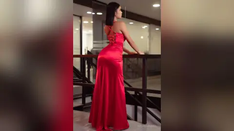 Elegant woman in a long red satin evening gown posing on a modern balcony at night.