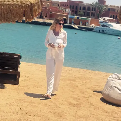 Woman in white linen outfit relaxing on a sunny beach by the turquoise ocean near a luxury resort.