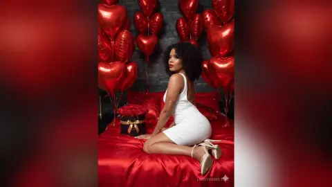 LoreyHamilton — Woman in white cocktail dress sitting on red bedding surrounded by red heart balloons for a romantic Valentine mood.
