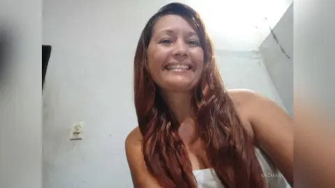 Smiling brunette woman with long wavy hair wearing a white top in a bright indoor setting.